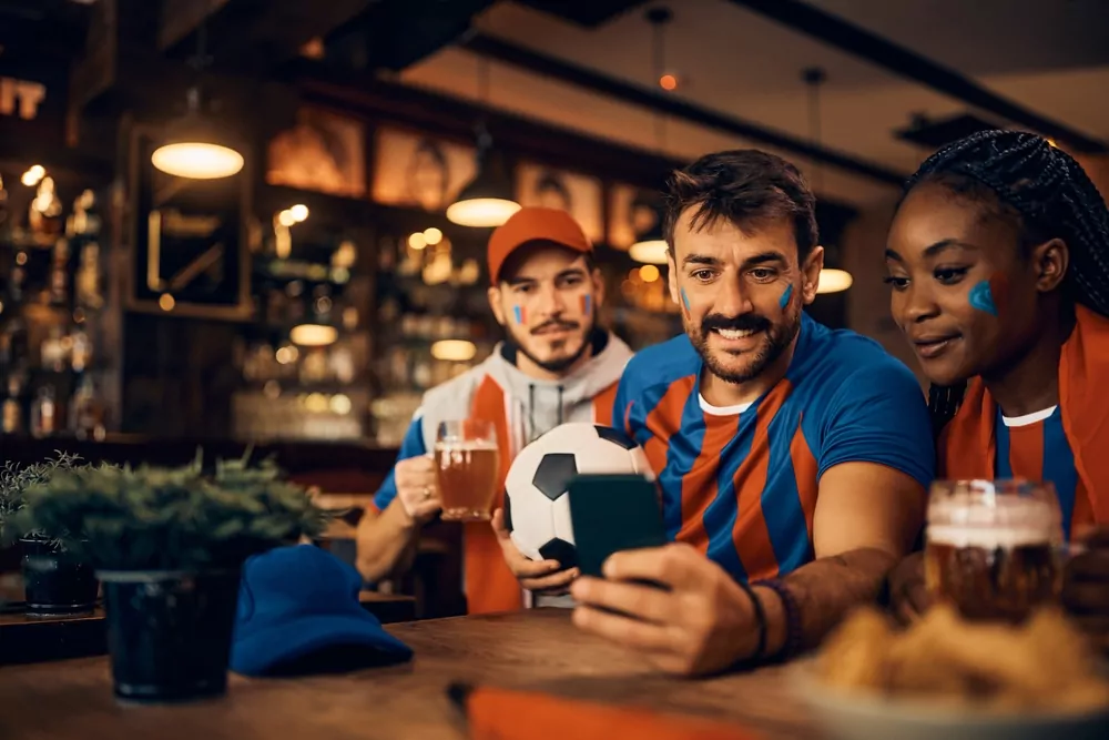 3 sports fans in orange and blue, at a bar using phone and sports technology